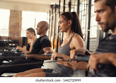 Group of people exercising in gym using rowing machine together. Side view of sportswoman doing exercise on rowing machine in center. Muscular girl and sporty men workout on training simulator at club - Powered by Shutterstock