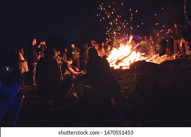 Group of people at the festival near camp fire with campfire song playing campfire games and eating camp fire grill, telling campfire stories near the fire with wood, flames in the nature at night