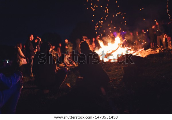 Group of people at the festival near camp fire with\
campfire song playing campfire games and eating camp fire grill,\
telling campfire stories near the fire with wood, flames in the\
nature at night