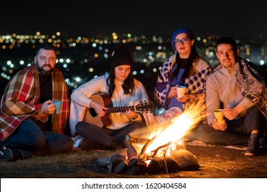 Group of people having fun sitting near bonfire outdoors at night playing guitar, singing songs and talking happily together.