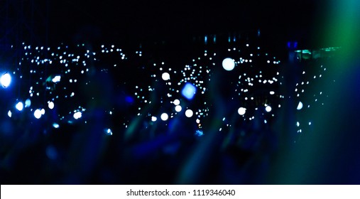 Group Of People Holding Cigarette Lighters And Mobile Phones At A Concert Crowd Of People Silhouettes With Their Hands Up. Dark Background, Smoke, Spotlights. Bright Lights