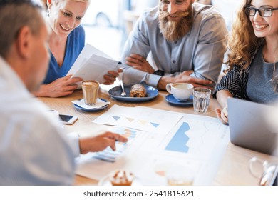 Group of people in a meeting, discussing charts and graphs. Smiling, diverse team collaborating over coffee. Business meeting with charts and teamwork. Diverse business people working together at cafe - Powered by Shutterstock