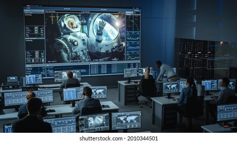 Group Of People In Mission Control Center Establish Successful Video Connection On A Big Screen With An Astronaut On Board Of A Space Station. Flight Control Scientists Sit In Front Of Computers.