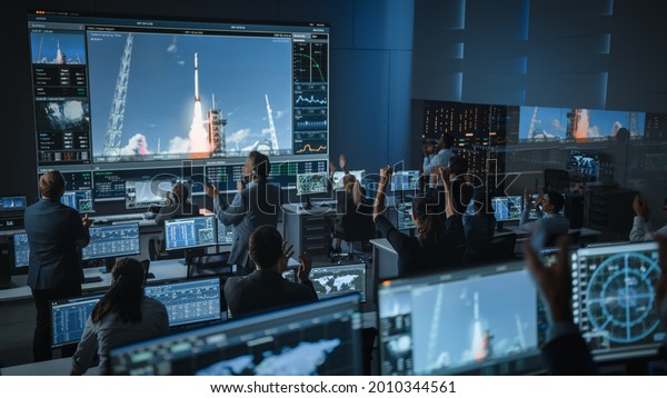 Group of People in Mission Control Center Witness\
Successful Space Rocket Launch. Flight Control Employees Sit in\
Front Computer Displays and Monitor the Crewed Mission. Team Stand\
Up and Clap Hands.