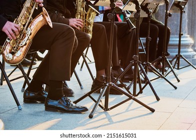 A Group Of People From A Musical Brass Band Who Are Sitting On Chairs And Playing Music, Jazz On Golden Brass Pipes, Sitting Outside In The Open Air. Sunshine On A Granite Column. Shiny Patent Shoes.