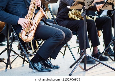 A Group Of People From A Musical Brass Band Who Are Sitting On Chairs And Playing Music, Jazz On Golden Brass Pipes, Sitting Outside In The Open Air. Shiny Patent Shoes.