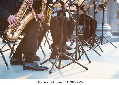 A Group Of People From A Musical Brass Band Who Are Sitting On Chairs And Playing Music, Jazz On Golden Brass Pipes, Sitting Outside In The Open Air. Sunshine On A Granite Column. Shiny Patent Shoes.