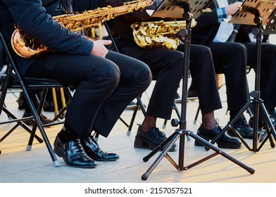 A Group Of People From A Musical Brass Band Who Are Sitting On Chairs And Playing Music, Jazz On Golden Brass Pipes, Sitting Outside In The Open Air. Shiny Patent Shoes.