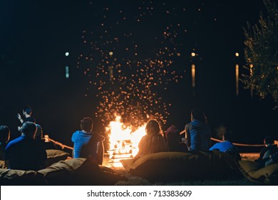 Group of people near camp fire with campfire song and campfire meals playing campfire games and eating camp fire grill, telling campfire stories near the fire with wood, flames in the nature at night.