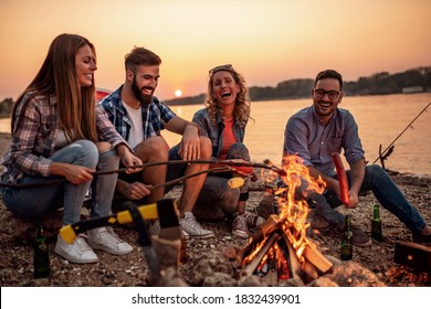 Group of people on camping sitting around campfire, drink beers, grilling sausages, having fun on beach at sunset.