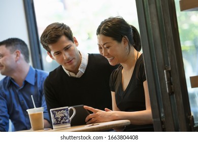 A group of people sitting around a table in a coffee shop. Looking at the screen of a digital tablet. Two men and a woman.
