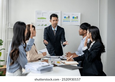 A group of people are sitting around a table with a man in a suit standing in front of them. The man is giving a presentation and the group of people are listening intently