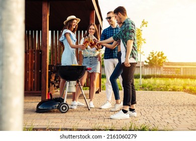 group of people stand around the grill, chatting, drinking and eating. Young friends are having a barbecue party at sunset in nature. Happy people barbecue outdoors, cook meat and drink drinks. - Shutterstock ID 2161060279