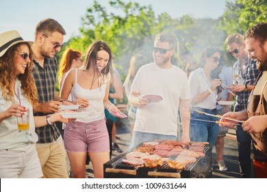 Group of people standing around grill, chatting, drinking and eating. 