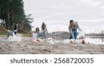 Group of people walking on beach of local lake and picking up trash left by others. Volunteers cleaning up nature from non organic materials. Wearing warm clothing and gloves for protection.