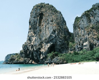 A group of people are walking on a beach near a large rock formation. The beach is empty and the sky is clear - Powered by Shutterstock