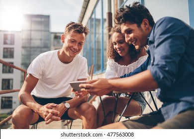 Group Of People Watching Video On The Mobile Phone While Sitting At Outdoor Cafe. Three Young Friends Sitting Outdoors And Looking At Smartphone.