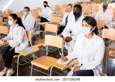 Group Of People In White Coats And Protective Masks Sitting In Conference Room Keeping Distance At Professional Training For Health Workers. Precautions During Mass Events In Coronavirus Pandemic