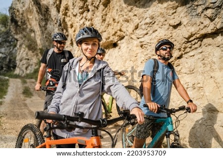 group of people woman men standing outdoor at the gorge cliff ridge at mountain range with their electric bikes bicycles in sunny summer day checking weather and location during ride taking a brake