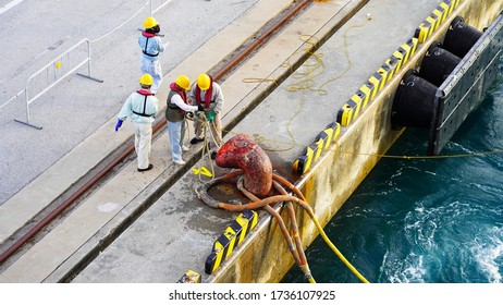  Group Of People Workers In Yellow Helmets And Life Jackets Moor A Cruise Ship In The Seaport, Many People Pull A Thick Rope Ashore. Workflow Security