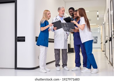 Group Photo Of Young Diverse Medics During Conversation With Senior Confident Doctor In Hospital Corridor Aisle. Medicine Concept