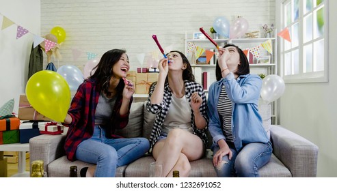 Group Portrait Of Cheerful Asian Friends Sitting On Sofa With Balloons And Toy In Hands Looking At Roof Blowing Whistles. Interior Of House Party On Background. Young Girls Having Fun Laughing.