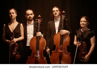 Group portrait of classical of string quartet looking at camera while standing on stage with warm top light against black curtains