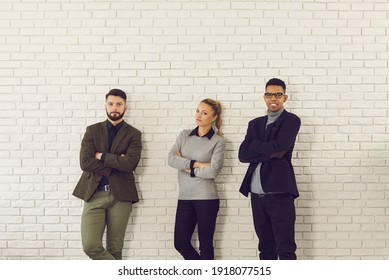 Group Portrait Of Confident Young Diverse Company Employees Or Managers. Team Of Successful Multiethnic Business People Looking At Camera, Standing Arms Folded Near White Brick Wall Of Loft Office