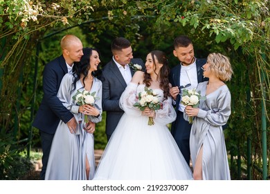 Group portrait of couples bride with groom and bridesmaids with groomsmen, posing in the garden near the arch, smiling looking at each other. Wedding day celebration, outdoors ceremony.
