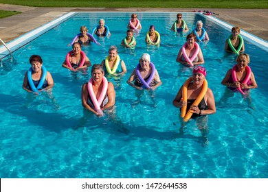 Group portrait of senior aqua gym class in outdoor pool. Senior ladies standing together in water with foam noodles around neck. 