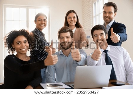Group portrait of smiling multiracial young businesspeople sit at desk in office show thumbs up together, happy diverse colleagues recommend good quality corporate service, acknowledgment concept
