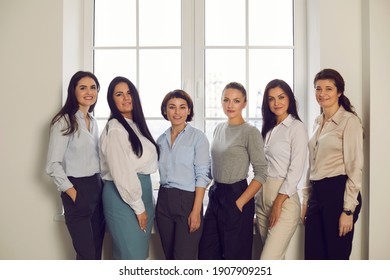 Group Portrait Of Successful Confident Stylish Women In Their 20s And 30s Standing Together By Office Window. Professional Team Of Happy Young Company Workers Or Business Partners Smiling At Camera