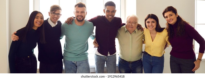 Group portrait of a team of cheerful confident diverse people embracing each other standing by the window together. Business partners, company employees or club members who are united by a common goal