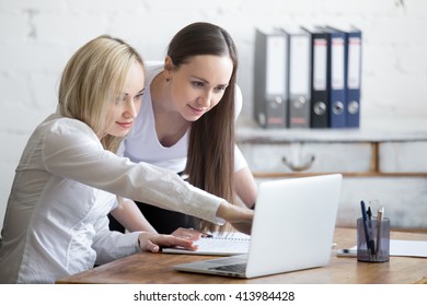 Group Portrait Of Two Beautiful Smiling Young Office Women Looking At Laptop Screen On Office Desk. Attractive Cheerful Business Ladies In Formal Wear Using Computer While Discussing Project