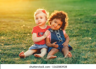 Group Portrait Of Two Cute Adorable Girls Toddlers Children Sitting Together. White Caucasian And Latin Hispanic Babies Hugging Outside In Park. Friendship And Best Friends Forever Concept. 