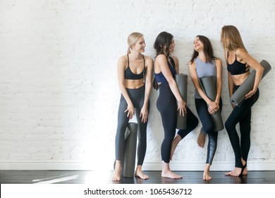 Group portrait of young sporty excited beautiful girls with exercise mats standing beside white wall laughing and talking together. Candid funny students waiting for class to start. Full length photo