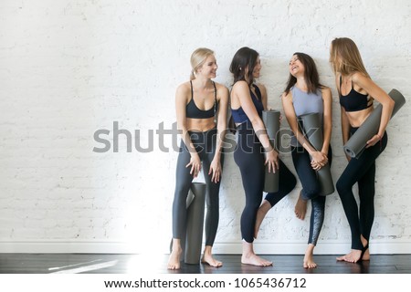 Group portrait of young sporty excited beautiful girls with exercise mats standing beside white wall laughing and talking together. Candid funny students waiting for class to start. Full length photo