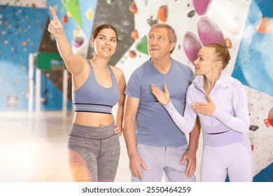 Group of positive friendly novice climbers, young women and elderly man, standing in training room against backdrop of bouldering wall, discussing with interest routes and techniques of climbing. - Powered by Shutterstock