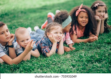 Group Of Positive Kids In Elementary School Age Lying On Green Grass In Park. The Concept Of Friendship, Childhood And Intercultural Communication.