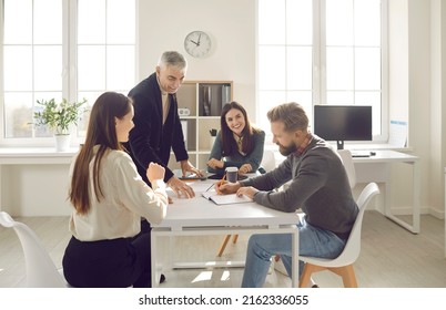 Group Of Positive People Dressed In Casual Clothes Discuss Plan For Cooperation On Common Project In Modern Coworking. People Write Their Ideas In Notebook Sitting At Table In Bright Office.