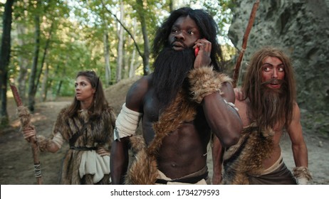 Group of primitive savages hunting animals in forest. African tribe leader using smartphone talking with friend on future gadget. Technology in prehistory.