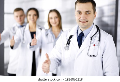 Group Of Professional Doctors Are Standing As A Team With Thumbs Up In A Hospital Office, Ready To Help Their Patients. Medical Help, Insurance In Health Care And Medicine Concept