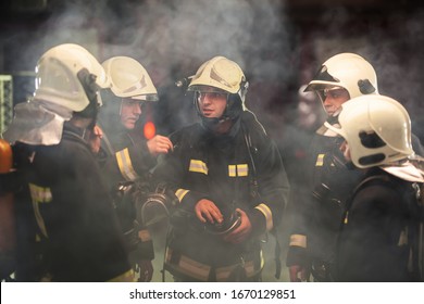 Group Of Professional Firemen Talking. Firefighters Wearing Uniforms And Protective Helmets. Smoke In The Background.