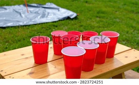 A group of red plastic cups are set up on a wooden picnic table, ready for a game or party. The background shows green grass and a tarp.