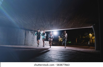 Group Of Runners Running Under A Bridge In City. Young Men And Women Jogging Together At Night.