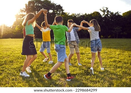 Group of school children playing games and having fun outdoors on a beautiful sunny summer day. Happy friends enjoying good weather, holding hands and dancing a round dance on green grass in the park