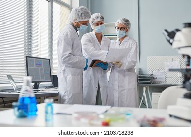 Group of scientists in protective wear holding document and discussing the results of analysis in team standing in the laboratory