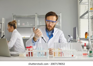 Group of scientists working in science laboratory. Serious young male pharma chemist or biotech company employee in white lab coat and protective glasses using pipette to transfer liquid in glass tube