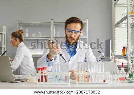 Group of scientists working in science laboratory. Serious young male pharma chemist or biotech company employee in white lab coat and protective glasses using pipette to transfer liquid in glass tube