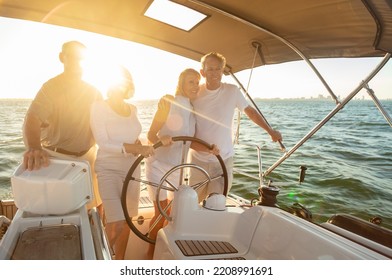 Group Of Senior Friends Enjoying Fun Summer Vacation Steering Luxury Yacht At Sunset With Females At The Helm